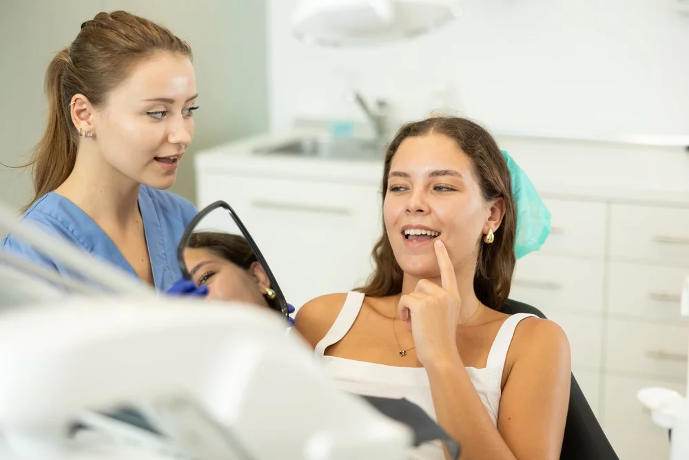 Happy girl admires her snow-white smile in the mirror. Female dentist helps her