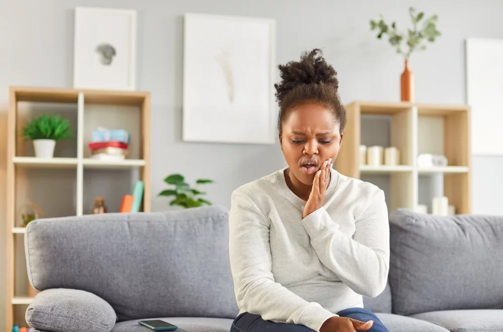Young woman sitting on the couch, holding side of cheek, clearly in tooth pain