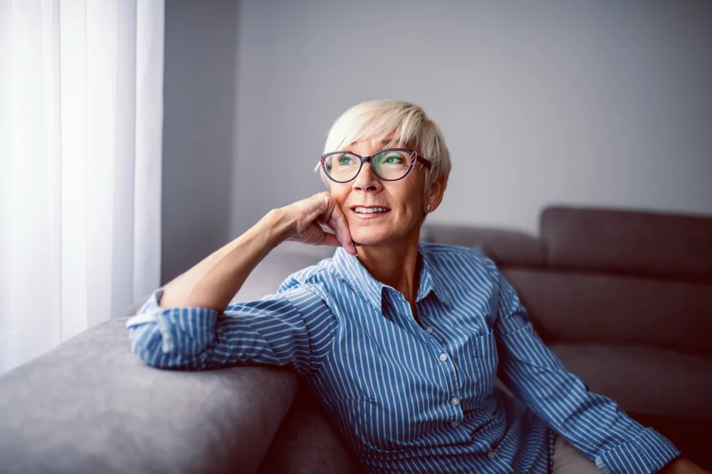 Older blonde woman sitting on a couch, hand on her face as she looks out the window